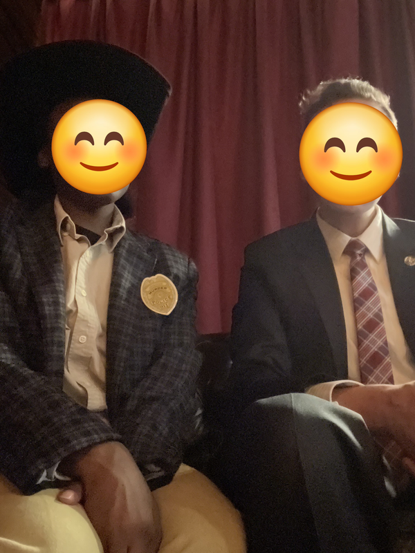 One person with a black tweed coat with white plaid, a black cowboy hat, khaki shirt, and fake police badge with a person in a black suit with a red with white stripe tie and a gold lapel pin; both are sitting in front of a red curtain