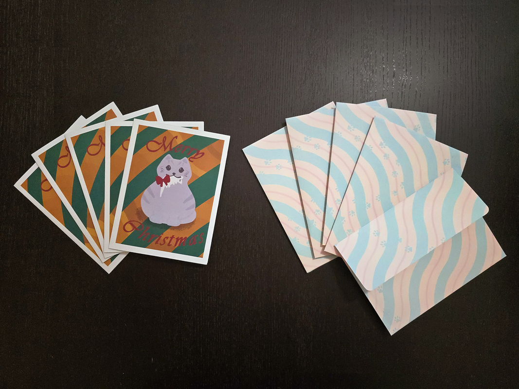 A small stack of Christmas cards, a small stack of envelopes with the back facing the camera, and a single envelope with the flap facing the camera. The cards have a round grey cat holding a dead mouse with a red bow on the mouse in the cat's mouth. The cat is standing in front of a gold and dark green striped background. The words "Merry Christmas" are printed on the card in maroon cursive. There is a white border around the edge of the card. The envelopes have parallel squiggly stripes diagonally covering the envelopes. The stripes are a repeating pattern of thick light green, medium thickness light yellow, thin light red, and medium thickness light yellow again. There is a path of paw-prints walking across the envelope in the same light green as the diagonal stripes. The envelope with the flap facing the camera is printed with the same pattern and reveals the flap to be a flat-edge flap with rounded corners.